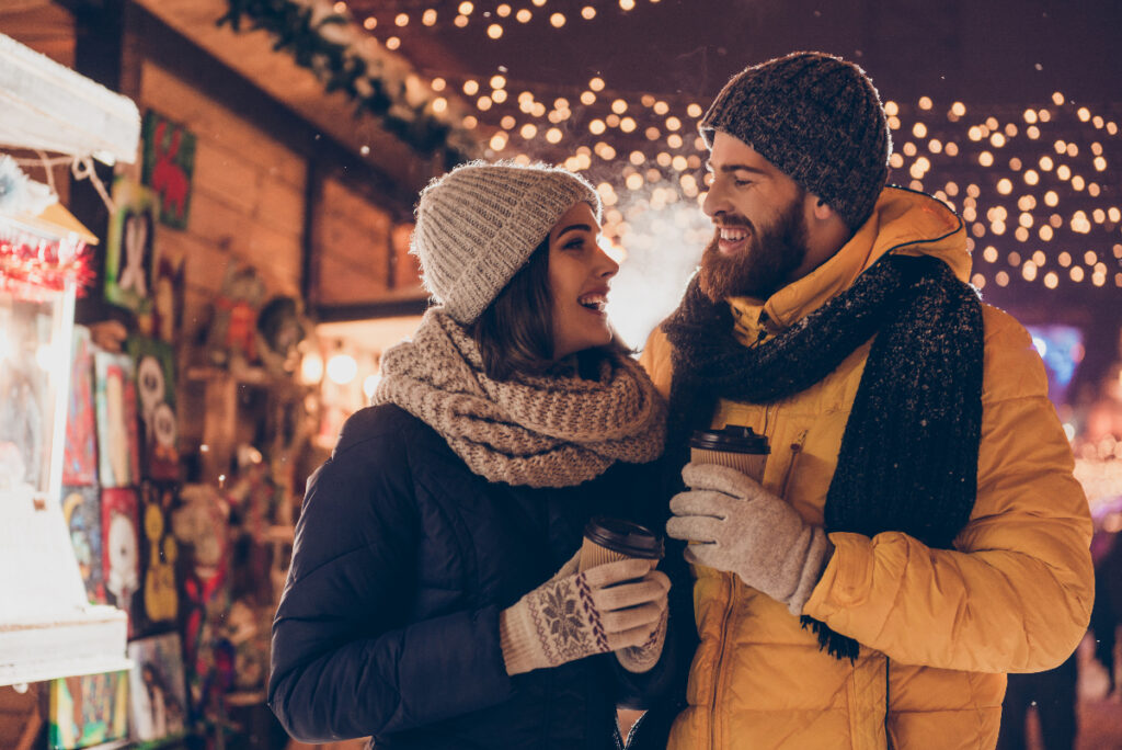 Having fun together at a christmas fairy. Young cheerful couple is having a walk with hot drinks, enjoying, dressed warm, looking at each other and laugh at Christmas market